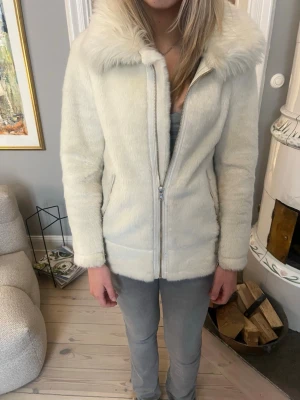 Vit pälsjacka med stor krage - Snygg vit pälsjacka med fluffig, bred krage och dragkedja framtill. Jackan har raka ärmar och två sidofickor. Materialet är mjukt och ger en lyxig känsla. Perfekt för dig som vill ha en stilren och trendig look.