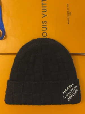  Louis Vuitton set - Säljer en svart stickad mössa från Louis Vuitton med rutmönster och uppvikt kant. På kanten finns vit brodyr med texten 'MARQUE L. VUITTON DÉPOSÉE'. Perfekt accessoar för dig som gillar exklusiv streetwear.