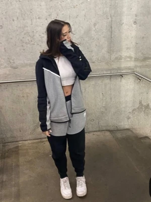 Nike  tech fleece  i svart och grå - Snygga mjukisbyxor från Nike i svart och ljusgrått med dragsko i midjan och sidofickor. Byxorna har en modern, avslappnad passform och är tillverkade i mjukt material som är perfekt för chill eller träning. Matchar med tillhörande hoodie för en komplett look.