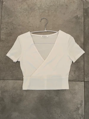 Vit omlottopp från Even&odd - Vit omlottopp med djup v-ringning, korta ärmar och croppad passform, från Even&odd💕 Köptes från Zalandos hemsida tillsammans med en likadan svart (Nypris 2st 220kr). Säljer för att den aldrig kommit till användning, så den är helt som ny!🩷 (Står ej för frakt och pris kan diskuteras)