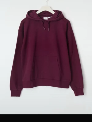 Vinröd hoodie med huva - En stilren vinröd hoodie med huva och dragsko. Hoodien har en klassisk känguruficka framtill och ribbade muddar vid ärmslut och nederkant. Perfekt för en avslappnad look och enkel att matcha med jeans eller joggers.