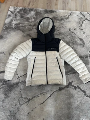 Vit och svart pufferjacka från McKinley - Snygg pufferjacka från Sail Racing i vitt med svart axelparti och huva. Jackan har två dragkedjeförsedda fickor framtill och quiltad design. Perfekt för kalla dagar med sin varma och lätta vaddering. Klassisk passform och sportig look.