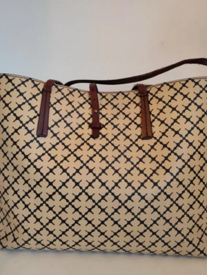 By Marlene Birger  - Stilren shoppingväska i beige med svart grafiskt mönster och bruna handtag. Väskan har en rymlig form och är perfekt för dig som vill ha plats för allt. Materialet känns tåligt och handtagen är i kontrasterande färg för en snygg look.