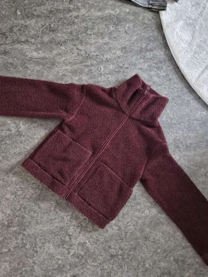 Vinröd teddyjacka med dragkedja - Mysig vinröd teddyjacka i fluffigt material med hög krage och dragkedja framtill. Jackan har två stora fickor och raka ärmar. Perfekt för kyliga dagar när du vill vara både varm och snygg. Endast använd 1 gång