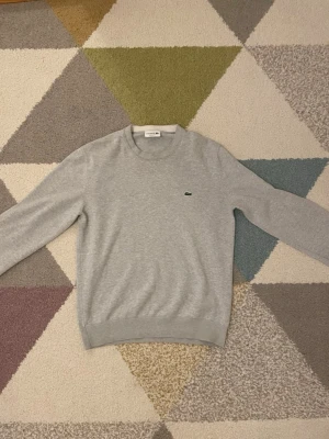 Grå sweatshirt från Lacoste - Klassisk ljusgrå sweatshirt från Lacoste med rund halsringning och diskret broderad krokodil-logga på bröstet. Tröjan har långa ärmar och ribbade muddar vid ärmslut och nederkant. Perfekt för en clean och avslappnad stil.