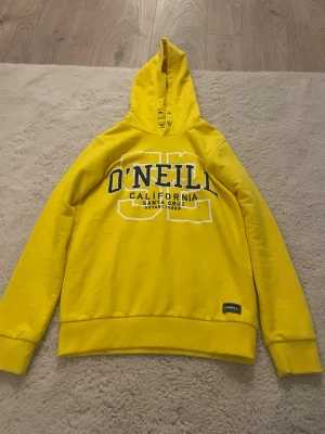 Gul hoodie från O'Neill - Snygg gul hoodie från O'Neill med tryck 'O'NEILL CALIFORNIA SANTA CRUZ' på bröstet. Klassisk modell med huva och ribbade muddar. Perfekt för en avslappnad och sportig stil. Materialet känns mjukt och bekvämt mot huden.