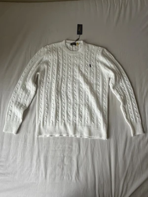 Vit kabelstickad tröja från Polo Ralph Lauren - Kabelstickad vit tröja från Polo Ralph Lauren med rund halsringning och den klassiska broderade loggan på bröstet. Tröjan har ribbade muddar vid ärmslut och nederkant. Perfekt för dig som gillar en clean och tidlös look.