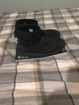 Svarta UGG tofflor i mocka - Säljer ett par svarta UGG tofflor i klassisk modell med låg skaft. Tillverkade i mjuk mocka med fårskinnsfoder för extra komfort. Perfekta för att hålla fötterna varma och sköna under kyliga dagar. Snygg och enkel design som passar till det mesta.