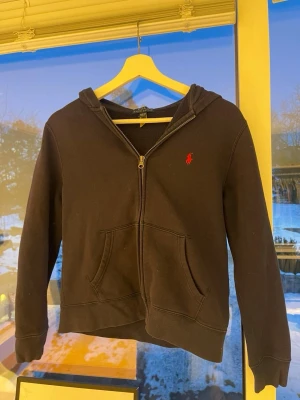 Marinblå  hoodie från Ralph Lauren - Marinblå hoodie med dragkedja från Ralph Lauren. Klassisk design med röd broderad logga på bröstet, två fickor framtill. Tillverkad i mjukt bomullsmaterial och har en skön huva. Perfekt för en avslappnad och stilren look. M (barnstorlek) 10-12 år, passar mig som är 13
