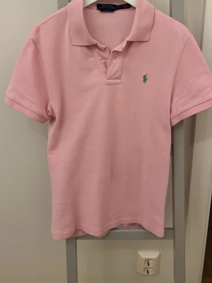 Rosa pikétröja från Ralph Lauren - Snygg ljusrosa pikétröja från Ralph Lauren med klassisk krage, korta ärmar och knappar framtill. Storlek L barn passar xs