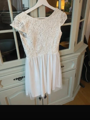 Vit spetsklänning med volang - Söt vit kortklänning med spets upptill och volangig kjol. Klänningen har korta ärmar, rund halsringning fram och djup v-ringning bak. Överdelen är dekorerad med broderad spets och kjolen är i ett lätt, flödande tyg.