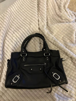 Svart handväska med silverdetaljer - Snygg svart handväska i slätt läder med coola silverfärgade metalldetaljer och dragkedjor. Väskan har dubbla handtag, dekorativa spännen och en framficka med dragkedja. Perfekt för dig som gillar en edgy och stilren look.