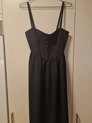 Svart klänning med smala axelband - En stilren svart klänning med smala axelband och figurnära överdel. Klänningen har en enkel, elegant design utan mönster och faller rakt ner från midjan. Perfekt för dig som gillar minimalistisk stil.