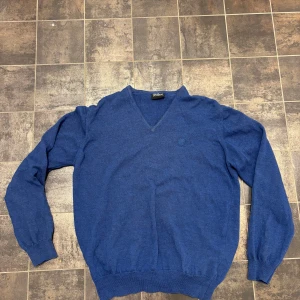 Blå V-ringad tröja Oscar Jacobson - Klassisk blå stickad tröja från Oscar Jacobson med V-ringning och ribbade muddar vid ärmslut och nederkant. Tröjan har en diskret logga broderad på bröstet och är tillverkad i mjukt material som känns skönt mot huden.                          Storlek: M men sitter som S