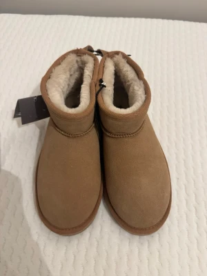 Bruna fårskinnstofflor med foder - Mysiga tofflor i brunt mockaliknande material med mjukt, ljust foder i fårskinnsimitation. Tofflorna har en enkel design med rund tå och låg ankel, perfekta för att hålla fötterna varma hemma. Dragsko upptill för bättre passform.