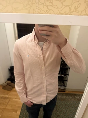 Rosa randig skjorta från Morris - Passar också S. Säljer en ljusrosa skjorta från Morris med tunna vita ränder. Skjortan har klassisk krage, knappar framtill och lång ärm. Tillverkad i mjuk bomull och har en liten broderad logga på bröstet. 