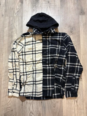 Hollister - Säljer en unik overshirt med svart och beige rutigt mönster, där skjortan är delad mitt på för en cool kontrasteffekt. Har en svart huva och knäppning framtill. Perfekt för dig som vill sticka ut med din stil. Passar M
