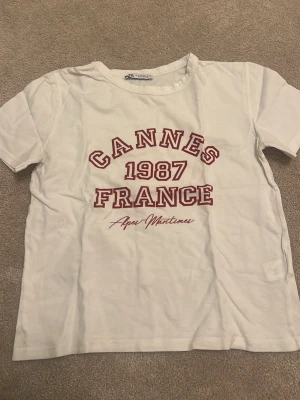 Zara t-shirt - Vit t-shirt från Aqua Maritime med röd text 'Cannes 1987 France' på bröstet. Klassisk rund hals och korta ärmar. Perfekt för dig som gillar retrovibbar och enkel stil.