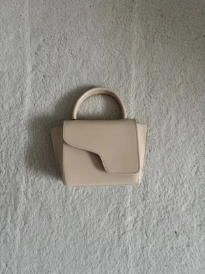Beige minihandväska med unik lockdesign - Stilren beige handväska i ministorlek med ett runt handtag och asymmetriskt lock. Väskan har hård struktur och är tillverkad i slätt konstläder. Perfekt för dig som gillar minimalistisk och modern design. väskan är från ATP och är endast använd fåtal gånger. Ordinarie pris är 3900kr