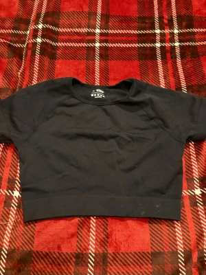 Svart croppad topp i syntet - En svart croppad topp med korta ärmar och rund halsringning. Tillverkad i stretchigt material med 92% polyamid och 8% elastan, vilket ger en tight och bekväm passform. Perfekt till träning eller som basplagg i garderoben.