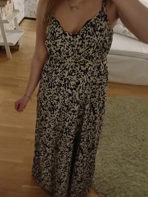 Svartvit långklänning med smala axelband - Långklänning med smala axelband och v-ringning. Klänningen har ett abstrakt svartvitt mönster och markerad midja. Tillverkad i ett lätt och mjukt tyg som faller snyggt längs kroppen. Perfekt för varma dagar och somriga tillfällen.