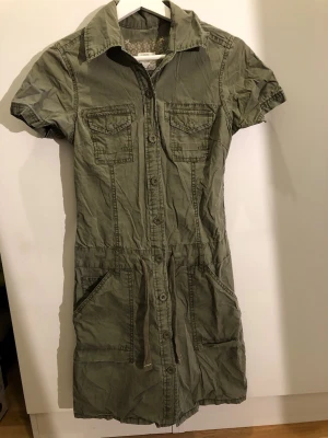 Grön skjortklänning med knappar - Snygg grön skjortklänning med korta ärmar, krage och knappar hela vägen framtill. Två bröstfickor och två stora fickor vid höfterna. Midjan har ett knytband för justerbar passform. 