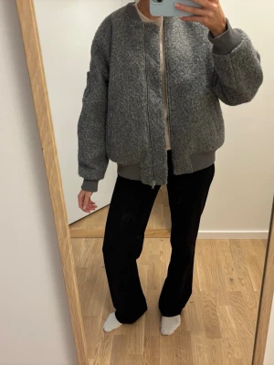 Grå bomberjacka i ullmix - Säljer en grå bomberjacka med grovstickad ullkänsla och ribbade muddar i ärmslut och nederkant. Jackan har dragkedja framtill, fickor på sidorna och en ärmficka med dragkedja. Oversized passform för en avslappnad look. Helt oanvänd!