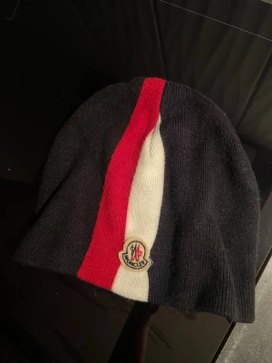 Moncler mössa med ränder - Snygg svart Moncler mössa i ribbstickad modell med en bred röd och vit rand framtill samt Moncler-logga. Mössan är mjuk och stretchig, perfekt för kyliga dagar. Klassisk design som passar till många stilar.