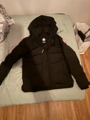 Svart dunjacka från Canada Goose - Snygg svart dunjacka från Canada Goose med huva och klassisk röd logotyp på ärmen. Jackan har dragkedja framtill, stora fickor och är vadderad med dun för extra värme. Perfekt för kalla dagar och har en modern, stilren look.