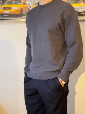 Stickad Crewneck  - En stilren mörkgrå tröja med rund hals och ribbade muddar. Tröjan är enkel och passar perfekt till alla typer av outfits. Tillverkad i mjuk bomull för skön komfort och avslappnad look.