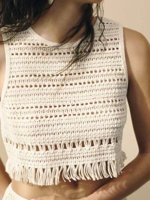 Beige virkad croppad topp - Beige virkad croppad linnetopp med fransar längst ner. Toppen är ärmlös och har ett snyggt hålmönster som ger en boho-vibe. Perfekt för sommardagar och temafester.