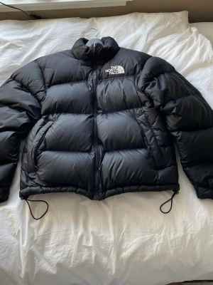 Svart dunjacka från The North Face - Säljer min svarta 1993 retro dunjacka från The North Face i storlek M. Den har en inbyggd luva,  två sidofickor med dragkedja och justerbar dragsko nedtill. Den är i värdigt bra skick och har använts omtänksamt. Perfekt för kalla vinterdagar. Ny pris ligger på 3 800 kr. Tveka inte att höra av dig om du har några funderingar