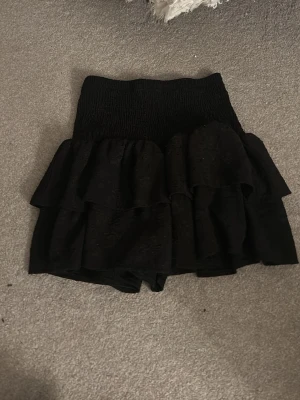 Svarta volangshorts med hög midja - Säljer ett par svarta shorts med hög midja och flera lager volanger. Shortsen har en smockad midja som ger en bekväm och stretchig passform. Perfekta för dig som vill ha en trendig och lekfull look.
