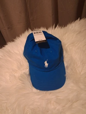 Blå keps från Polo Ralph Lauren - Snygg blå keps från Polo Ralph Lauren med klassisk vit broderad logga framtill och 'POLO' broderat bak. Justerbart spänne i metall baktill för perfekt passform. Tillverkad i bomull för en skön och luftig känsla.