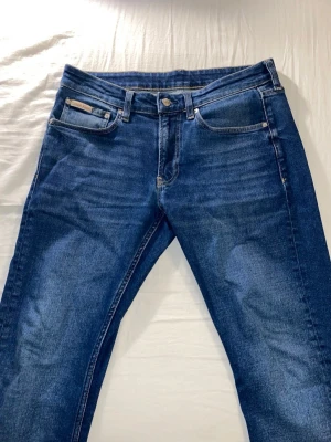 Mörkblå jeans från Calvin Klein - Snygga mörkblå jeans från Calvin Klein med klassisk femficksmodell och raka ben. Jeansen har subtila slitningar och kontrastsömmar som ger en modern look. Tillverkade i mjuk och slitstark denim som passar perfekt till vardagsstilen.