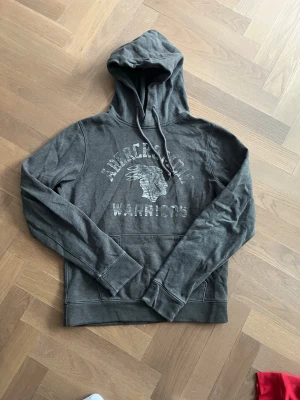 Abercrombie & Fitch hoodie - Snygg mörkgrå hoodie med tryck framtill från Abercrombie & Fitch. Ruggigt coolt tryck som ger hela plagget en archive känsla. Strl M. 