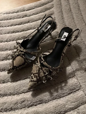 Ormskinnsmönstrade klackskor med remmar - Snygga pumps med spetsig tå och ormskinnsmönster i beige och svart. Skorna har tunna remmar över foten och runt vristen samt en smal klack. Perfekta för dig som vill sticka ut med en trendig look. Har endast testat dom hemma 1 gång sen inte användt dom