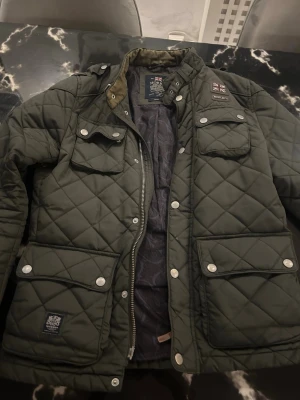 Olivgrön quiltad jacka från Morris - Snygg olivgrön quiltad jacka från Morris med fyra framfickor, silverfärgade knappar och dragkedja. Jackan har ståkrage med spänne och patch med brittisk flagga på bröstet. Perfekt för dig som gillar klassisk men trendig stil.