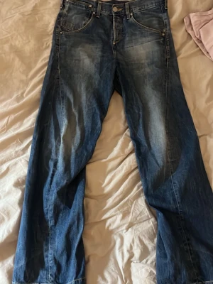 Blå Levi's jeans med vintage look - Säljer ett par blå Levi's jeans med snyggt slitna detaljer och klassisk femficksmodell. Jeansen har raka ben och en cool vintage tvätt. Bakfickan har den ikoniska Levi's-loggan och det finns ett justerbart bälte bak i midjan.