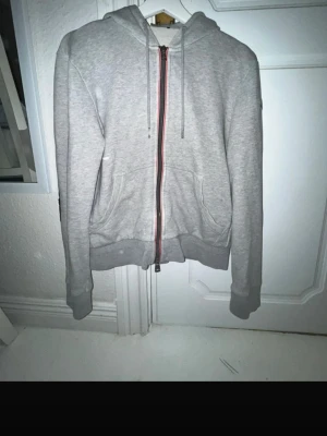 Moncler kofta - Snygg grå hoodie från Moncler med dragkedja framtill och två fickor. Klassisk huva med dragsko och Moncler-logga på ärmen. Tillverkad i mjukt material som känns skönt mot huden. Perfekt för en avslappnad och trendig look. Storlek Herr Xs. Använt i bra och sparsamt skick. Bara 700kr! Tror den är äkta.
