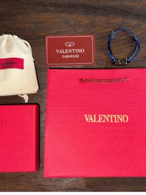Valentino blått armband med logga - Snyggt armband från Valentino Garavani i marinblått tygband med justerbar knytning och metallspänne i form av Valentinos ikoniska V-logga. Kommer med originalpåse, ask och presentpåse. Perfekt accessoar för dig som gillar exklusiva detaljer. (Allt i Bilden medföljer)