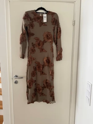 Beige långklänning med blommor - Snygg beige långklänning med brunt blommönster och långa ärmar. Klänningen har en kroppsnära passform och är tillverkad i ett tunt, stretchigt material som känns bekvämt. Perfekt för dig som vill sticka ut med ett unikt mönster. Nypris 1 000kr