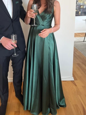 Grön långklänning i satin - Elegant grön långklänning i glansigt satinmaterial med omlott över bysten och v-ringad hals. Klänningen har tunna axelband som knyts vid axeln och ett svepande fall som ger en lyxig känsla. Perfekt för speciella tillfällen. Endast använd en gång. Från Jolie bridral. Ny pris 2500kr