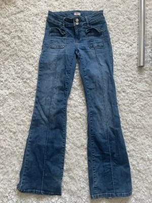 Blå bootcut jeans från Nelly - Säljer ett par blå bootcut jeans från Nelly med låg midja och dubbla knappar. Jeansen har stora fickor framtill och markerade sömmar längs benen. Tillverkade i klassiskt denimtyg, perfekta för en trendig och avslappnad look.
