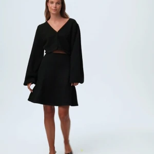 Svart stickad klänning med v-ringning - Snygg svart klänning med v-ringad topp och långa ärmar. Klänningen har en croppad topp med knappar och en kjol i normal passform. Perfekt för en stilren look.