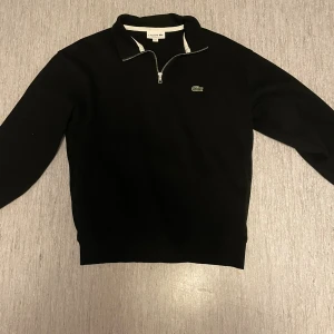 Svart half zip tröja från Lacoste - Svart långärmad tröja från Lacoste med klassisk krokodillogga på bröstet. Tröjan har en snygg half zip-dragkedja och vit kant vid kragen. Classic fit och tillverkad i mjuk bomull som känns skön mot huden. Perfekt för dig som gillar stilrena plagg. Hör av er vid frågor! 