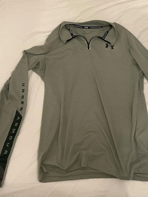 Grå långärmad träningströja Under Armour - Snygg grå långärmad träningströja från Under Armour med halv dragkedja och svart logga på bröstet. Ärmen har en svart detalj med texten UNDER ARMOUR. Perfekt för träning eller löpning, tillverkad i ett lätt och ventilerande material.