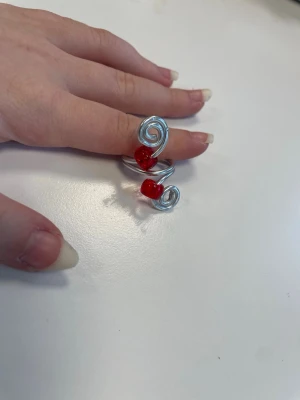 Silvrig ring med röda pärlor - Unik handgjord ring i silvrig metalltråd med två röda pärlor. Ringen har en öppen design med snygga spiraler på båda sidor och de röda pärlorna sitter i mitten. Perfekt för dig som gillar statement-smycken och vill sticka ut.