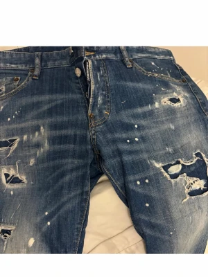 Blå slitna jeans från Dsquared2 - Säljer ett par blå jeans från Dsquared2 med distressed detaljer, slitningar och färgstänk för en riktigt edgy look. Klassisk femficksmodell med knappgylf och röd Dsquared2-lapp bak. Perfekta för dig som gillar streetwear och statement-plagg.