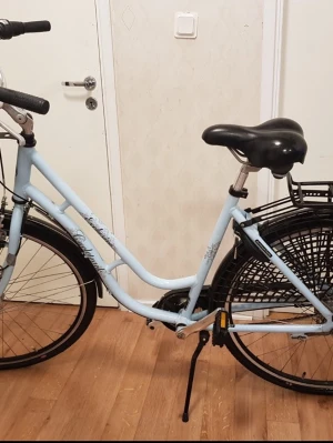 Ljusblå damcykel från Cresent - Säljer en ljusblå damcykel från Crescent med klassisk ram, svart sadel och korg framtill. Cykeln har kedjeskydd, pakethållare och reflexer. Stänkskärmar och handtag i svart. Perfekt för dig som vill ha en snygg och praktisk cykel.